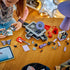 LEGO Olivia Rodrigo Secret Storage pieces in black, gray, lavender and red cover a wood table as a builder assembles the music case beside digital instructions.