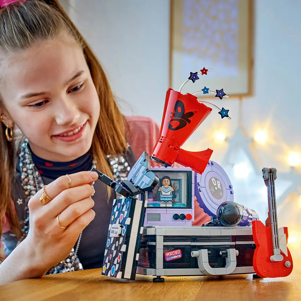 LEGO Olivia Rodrigo Secret Storage in black, gray, lavender and red bricks forms a music case display as a smiling builder adjusts the butterfly panel at a wood table.