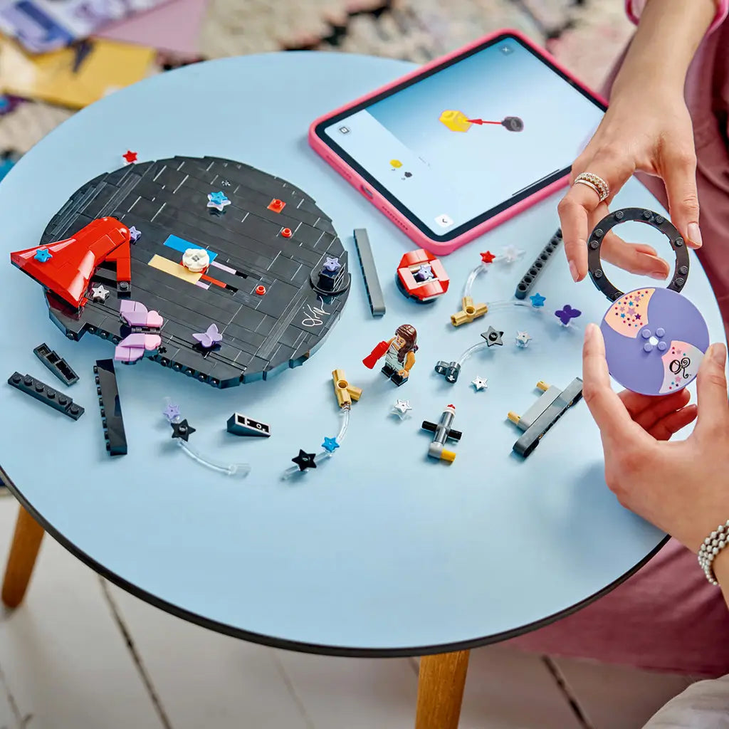 LEGO Olivia Rodrigo Vinyl set in black, purple and red bricks is being assembled on a blue table as hands hold the purple disc beside loose pieces and a tablet.