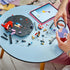 LEGO Olivia Rodrigo Vinyl set in black, purple and red bricks is being assembled on a blue table as hands hold the purple disc beside loose pieces and a tablet.