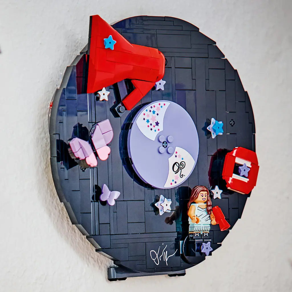 LEGO Olivia Rodrigo Vinyl display in black, purple and red bricks is mounted on a light wall, featuring a purple disc, red megaphone, butterflies, stars and minifigure.