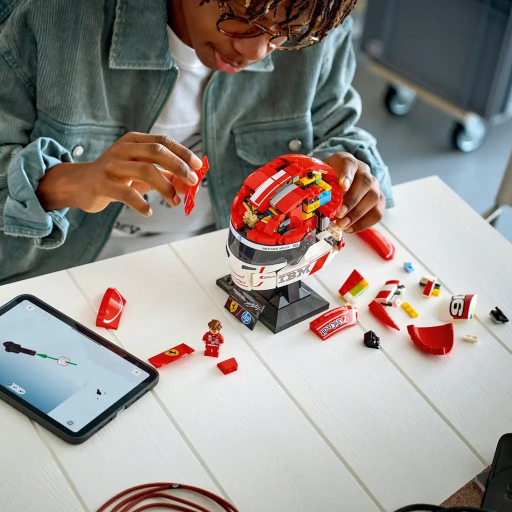 LEGO Ferrari Charles Leclerc helmet building set features red, white and black plastic brick components forming a Formula 1 racing helmet, with a builder assembling panels beside digital instructions on a tabletop.