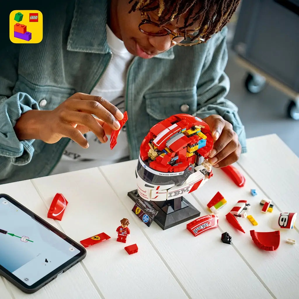 LEGO Ferrari Charles Leclerc helmet model features red, white and black glossy plastic brick construction shaped as a Formula 1 racing helmet on a display stand, shown during assembly with digital instructions and parts on a table.