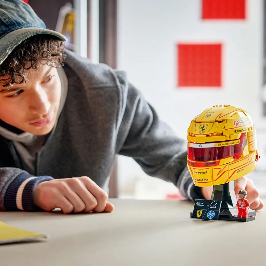 LEGO Ferrari Formula 1 helmet display features metallic yellow and red plastic panels with a tinted visor, Lewis Hamilton nameplate and Ferrari minifigure showcased on a stand in a bright indoor setting.
