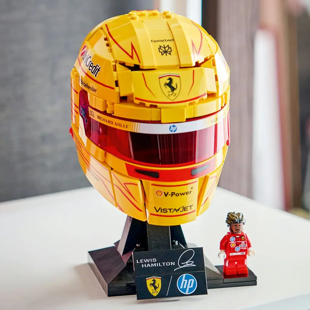 LEGO Ferrari Formula 1 helmet collectible features metallic yellow and red plastic surfaces, a reflective visor, sponsor markings and Lewis Hamilton Ferrari minifigure displayed on a stand indoors.
