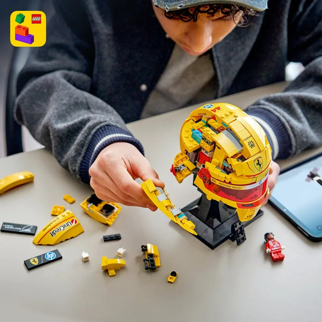 LEGO Ferrari Formula 1 helmet model features metallic yellow and red plastic panels with racing graphics, a tinted visor and Lewis Hamilton Ferrari minifigure displayed on a stand in a modern room.
