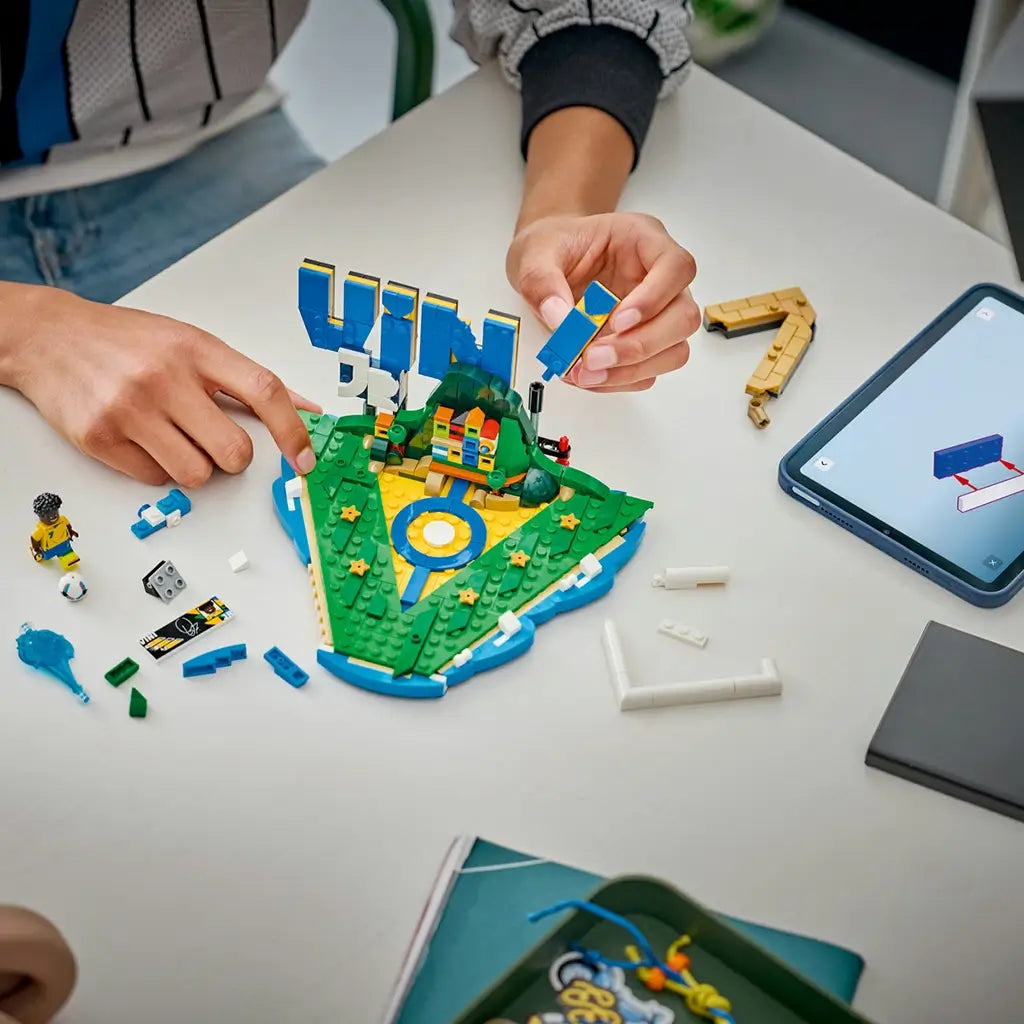 LEGO Editions Vini Jr. Soccer Highlights building set shows blue, yellow, green and white plastic parts, a football player minifigure and interactive display sections being assembled on a tabletop beside digital instructions.
