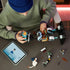 LEGO Fortnite Kit set 77081 in blue, black and gray plastic bricks is assembled by a builder at a table, with robot sections, loose pieces and digital instructions nearby.