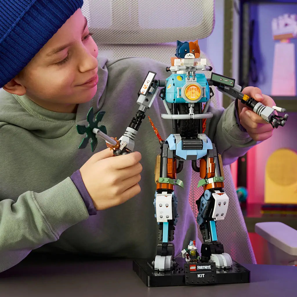 LEGO Fortnite Kit robot in blue, black, gray and brown plastic bricks is posed by a builder holding its articulated arm and green pickaxe in a colorful gaming room.