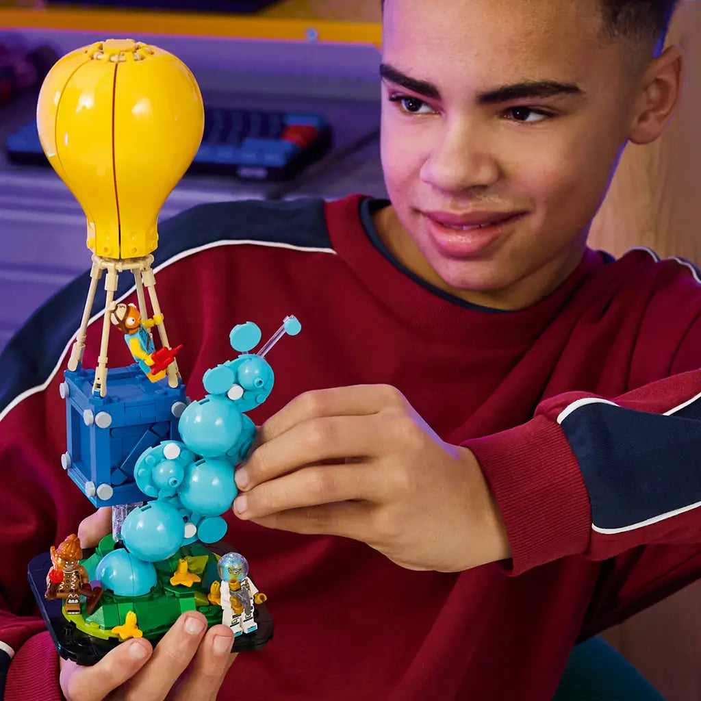 LEGO Fortnite Supply Drop set in yellow, blue and turquoise plastic bricks is held by a builder, featuring a balloon crate, bubble effect, green base and three figures.
