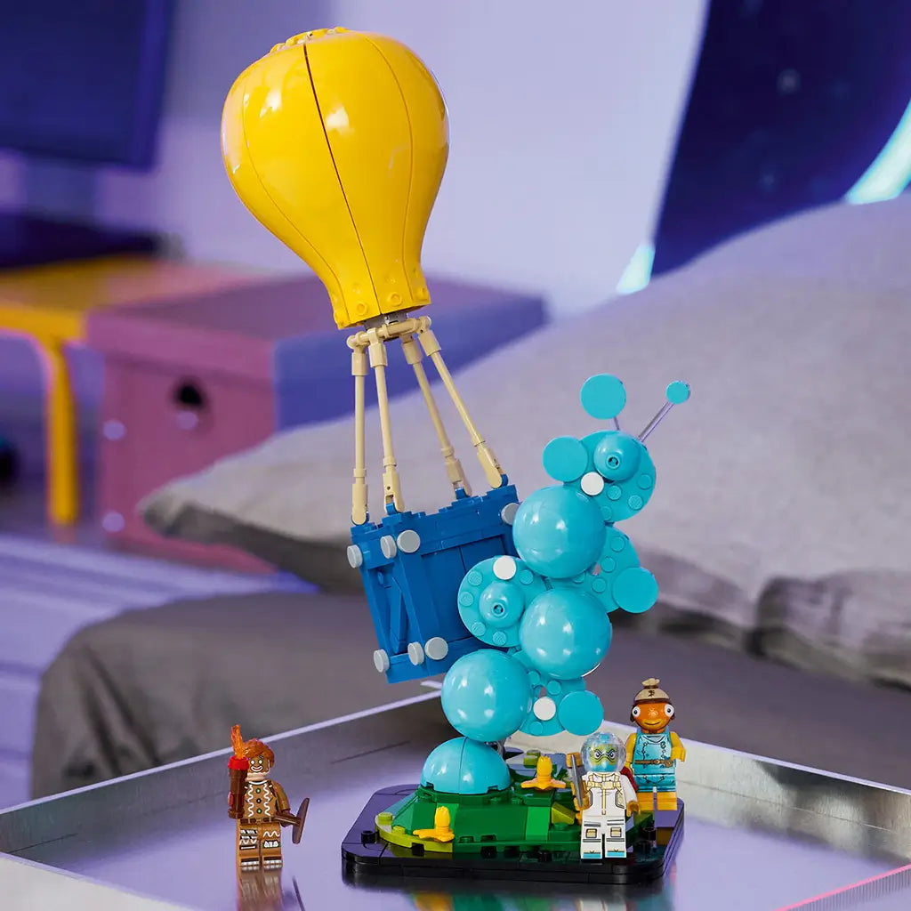 LEGO Fortnite Supply Drop set in yellow, blue and turquoise plastic bricks, featuring a balloon crate, bubble effect, green terrain base and three figures in a gaming room.