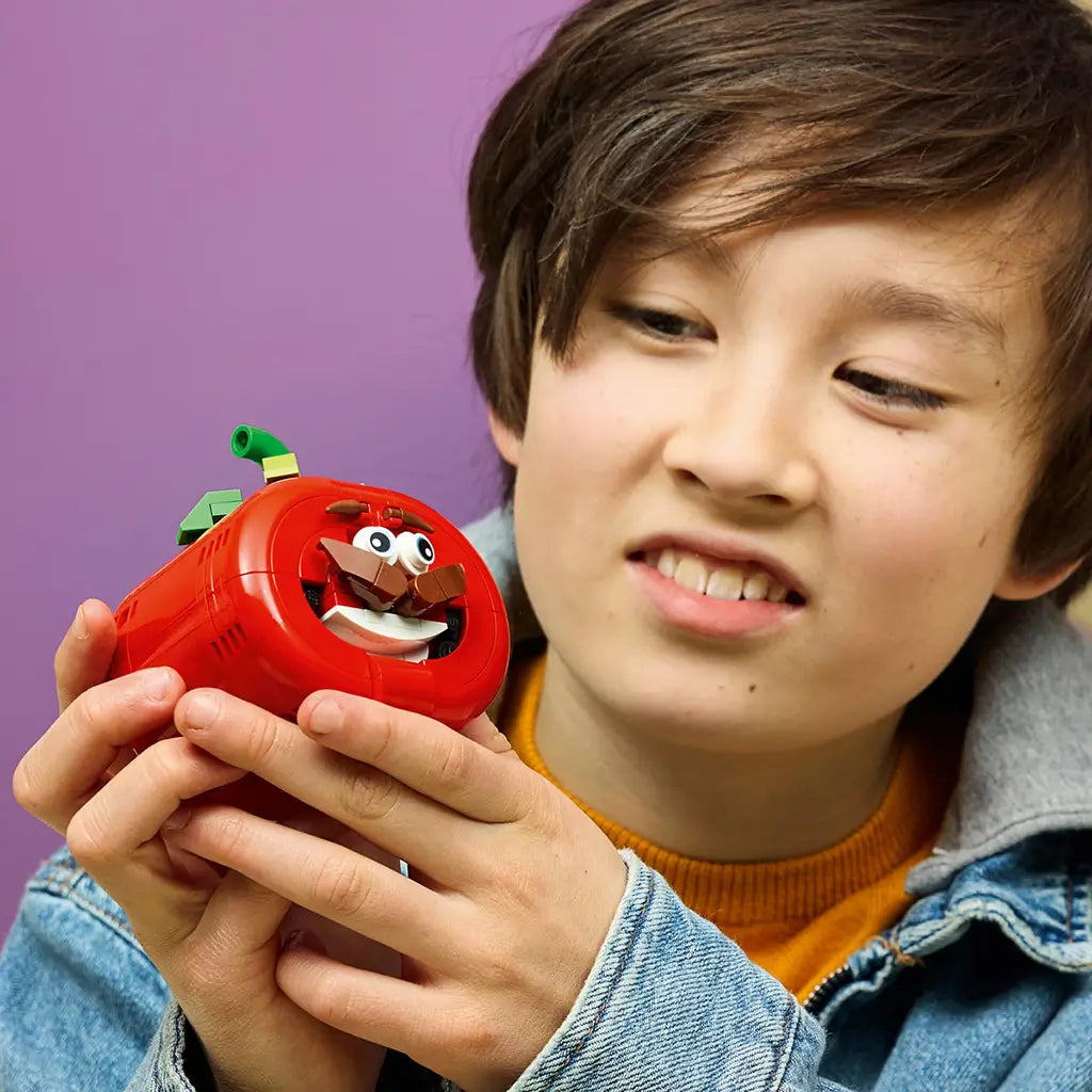 LEGO Fortnite Tomatohead set in red, green and brown plastic bricks is held by a child, showing the rounded tomato form, wide eyes, mustache, white grin and leafy stem.