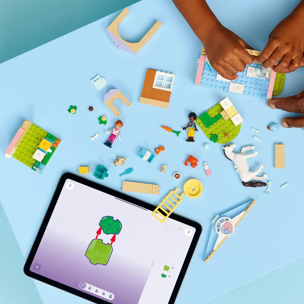 LEGO Friends animal vet clinic building activity on a light blue table, pastel plastic bricks, animals, and tools spread out, showing a child assembling the clinic structure while following digital building instructions on a tablet.