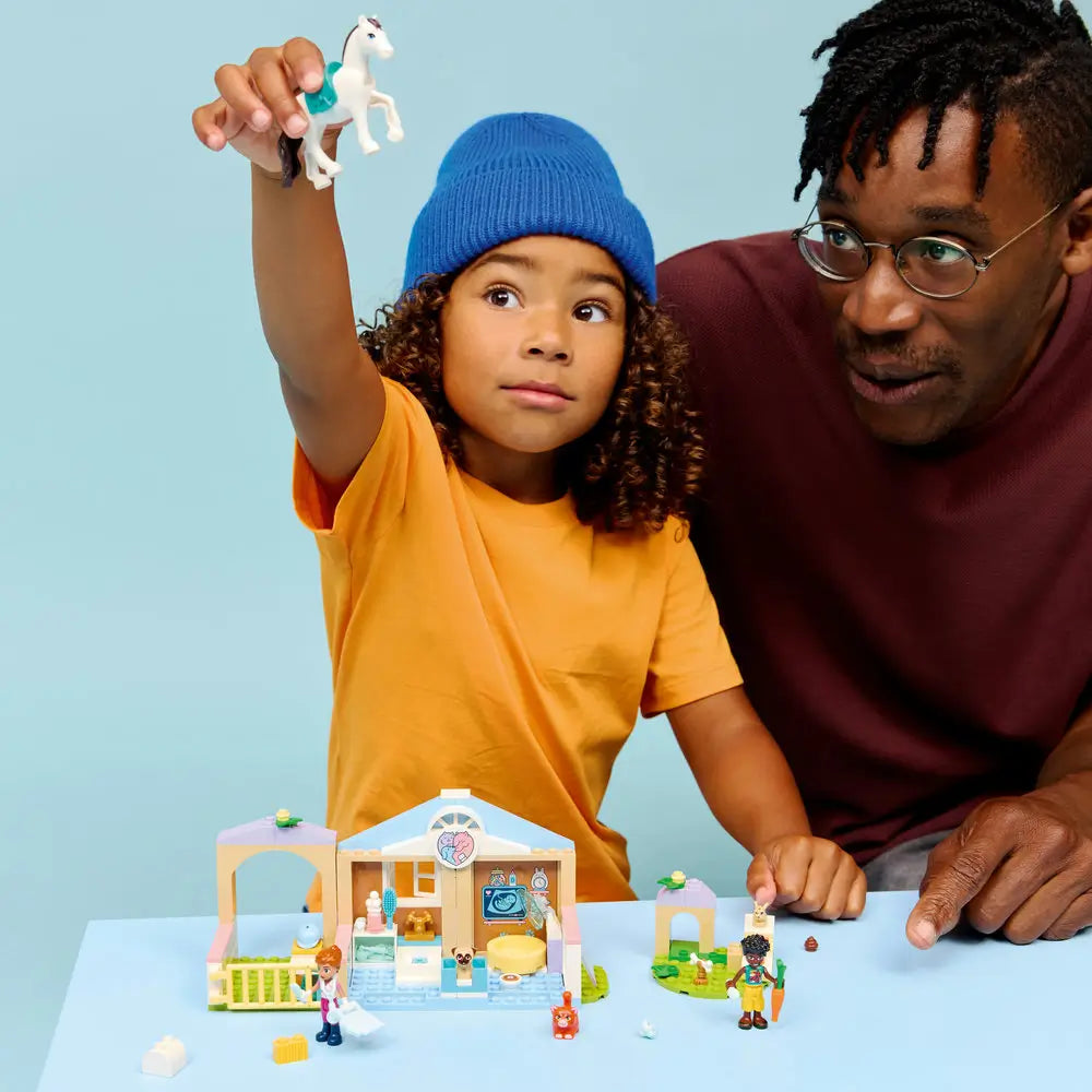 LEGO Friends animal vet clinic playset used during shared play, pastel clinic building with animals and accessories, shown with a child and adult interacting and pretending to care for pets in a bright indoor setting.