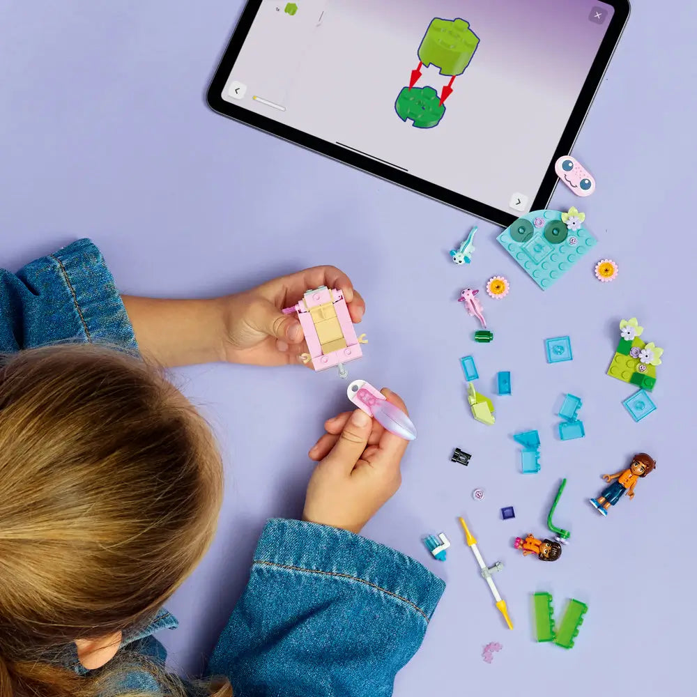 LEGO Friends lifestyle image showing a child building an axolotl themed boat and pond from pastel plastic bricks, connecting parts while figures, animals, and accessories are arranged for imaginative outdoor play.