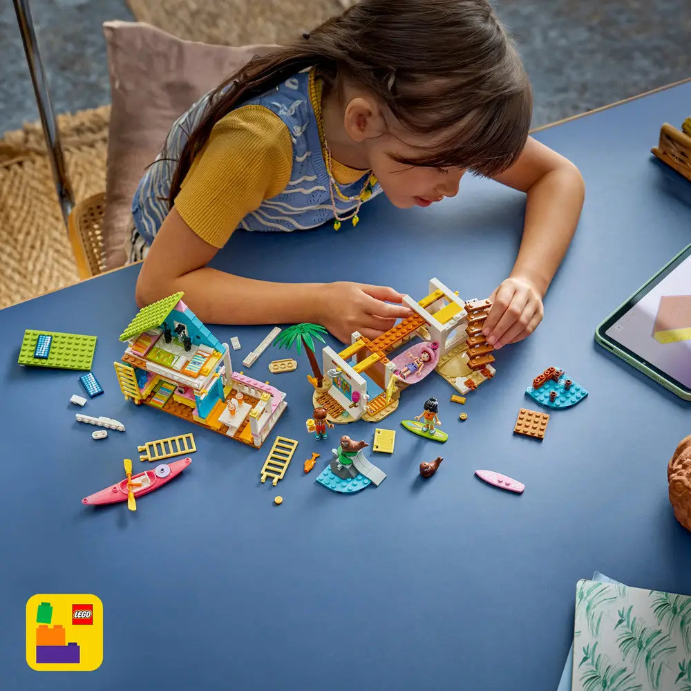 LEGO Friends beach house building set shown disassembled in colorful plastic pieces, raised house sections, dock, palm tree, and water elements, illustrating modular build stages and play features against a white studio background.