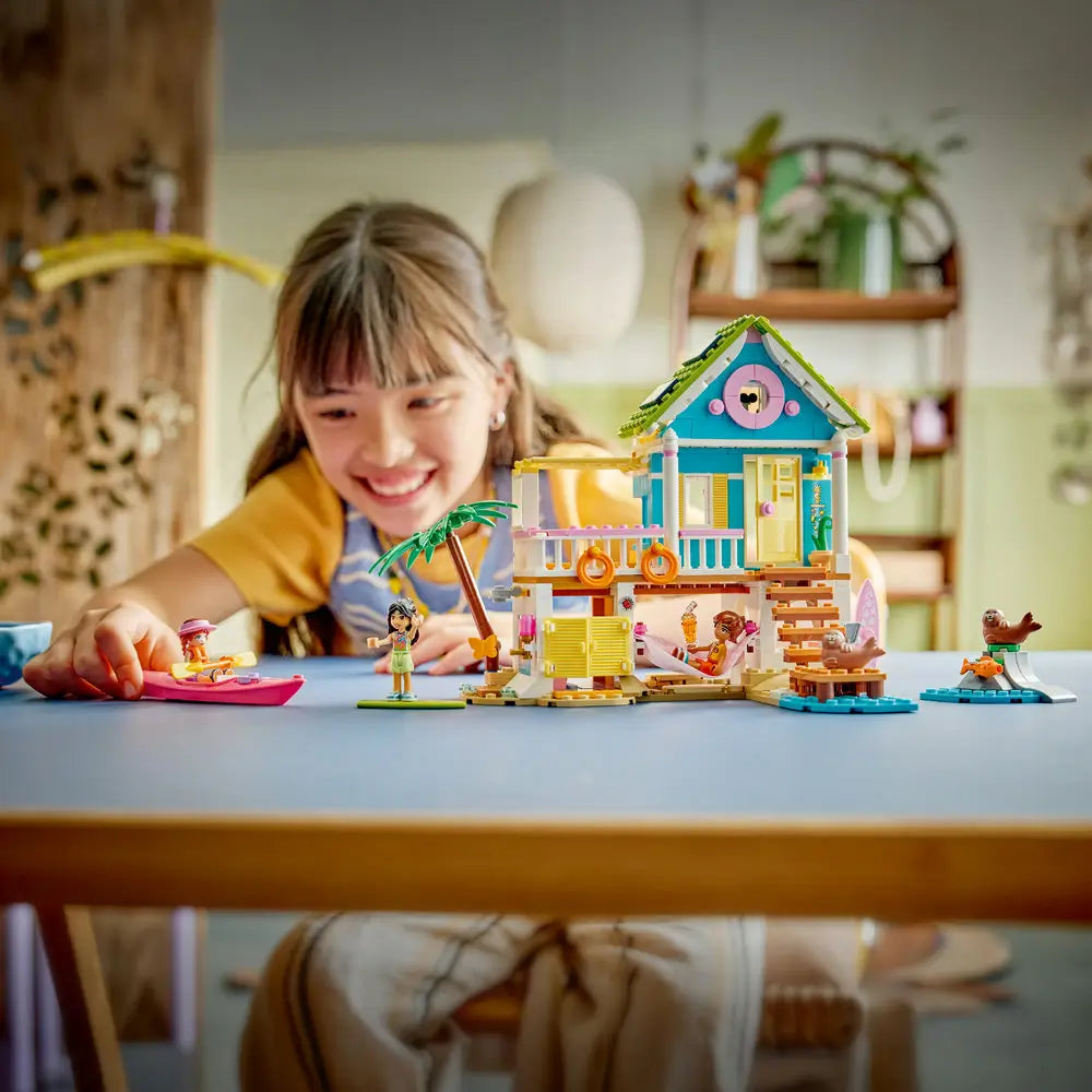 LEGO Friends beach house playset displayed during shared play, turquoise and yellow stilt house with balcony, palm tree, kayak, surfboards, and seal figures, shown with an adult and child enjoying imaginative indoor play.