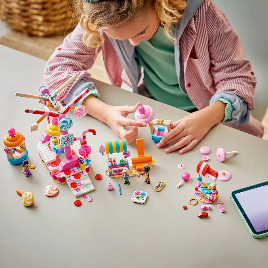 LEGO Friends Candy & Cupcake Ferris Wheel 42700 in pink plastic is assembled by a child at a table, with ride parts, candy stalls, mini-dolls, accessories, and a tablet nearby.
