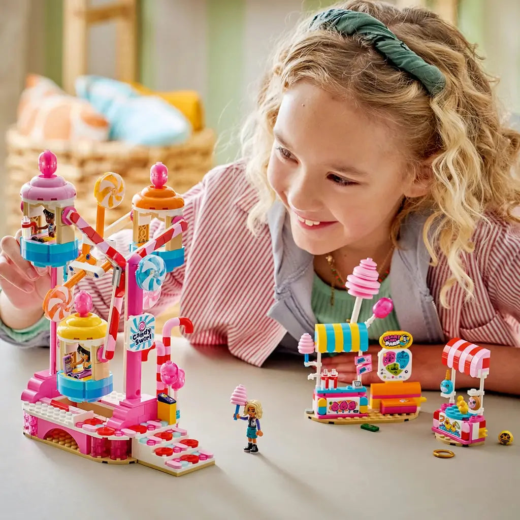 LEGO Friends Candy & Cupcake Ferris Wheel 42700 playset in pink and blue plastic is explored by a smiling child, with candy ride, snack stands, and mini-dolls arranged on a table.