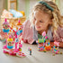 LEGO Friends Candy & Cupcake Ferris Wheel 42700 playset in pink and blue plastic is explored by a smiling child, with candy ride, snack stands, and mini-dolls arranged on a table.