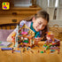 LEGO Friends Cozy Fall Forest Cabin playset opens to detailed tan and brown rooms as a child builds at a table surrounded by figures, autumn trees, bicycle and loose bricks.