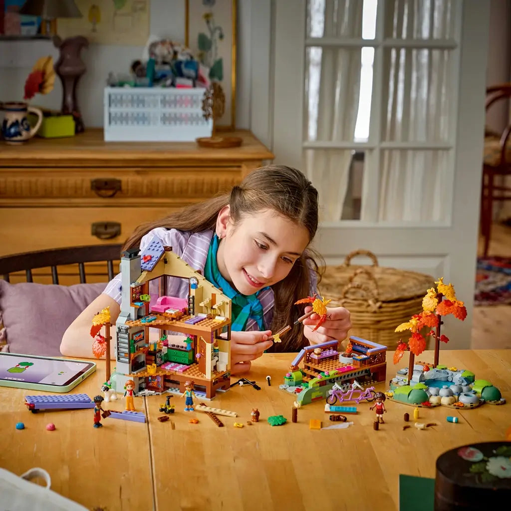 LEGO Friends Cozy Fall Forest Cabin building scene shows a child assembling the open brown and tan lodge at a table with figures, autumn trees, bicycle and scattered pieces.