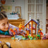 LEGO Friends Cozy Fall Forest Cabin in brown, tan and purple bricks is arranged on a table as a child plays with figures, autumn trees, rocky stream, bicycle and woodland details.