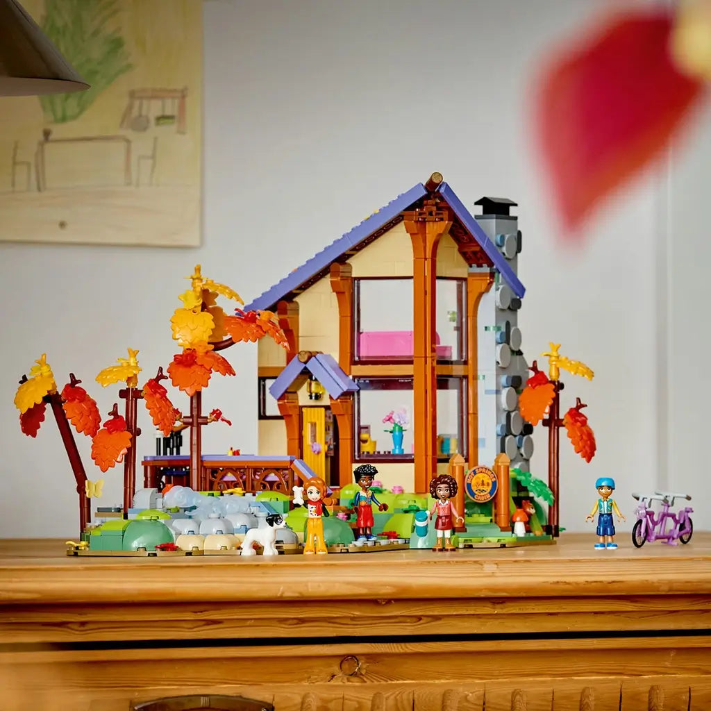 LEGO Friends Cozy Fall Forest Cabin display features a tall brown and tan lodge with purple roof, gray chimney, orange autumn trees, rocky stream, figures and bicycle on a wood surface.