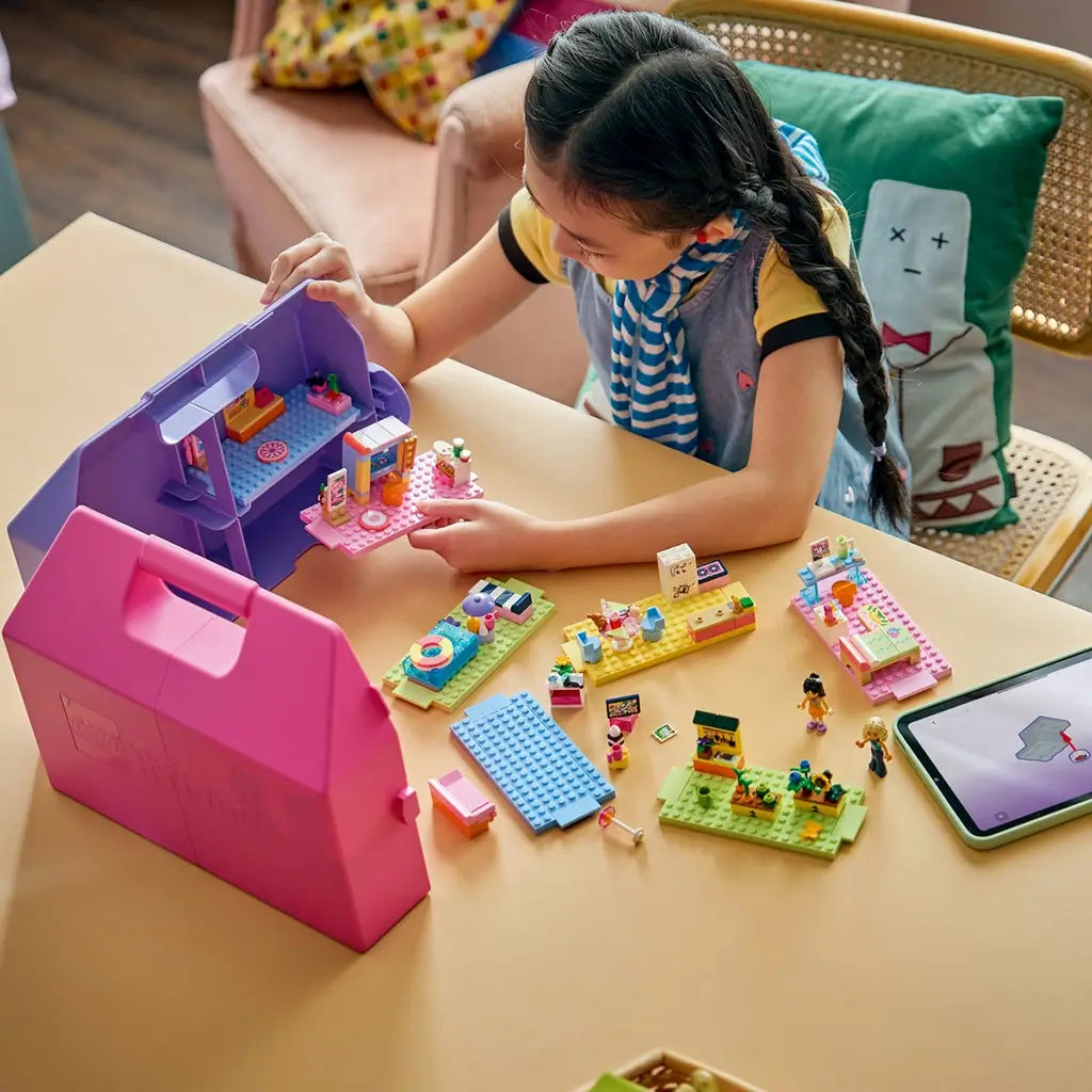 LEGO Friends Creative Dollhouse Suitcase in pink and purple plastic surrounds colorful room builds as a child assembles pieces beside a tablet on a home table.