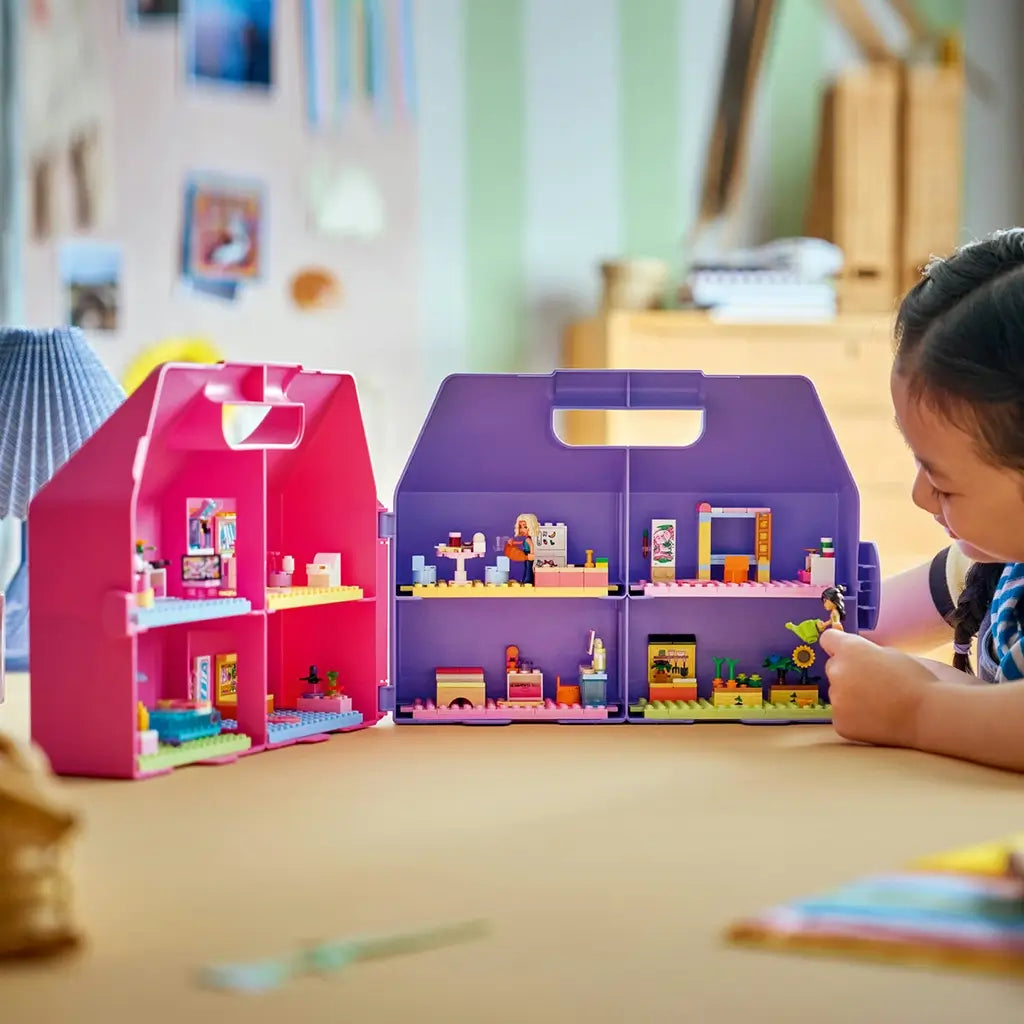 LEGO Friends dollhouse suitcase in pink and purple plastic opens into four furnished rooms while a child plays with a mini-doll and plant pieces at a home table.