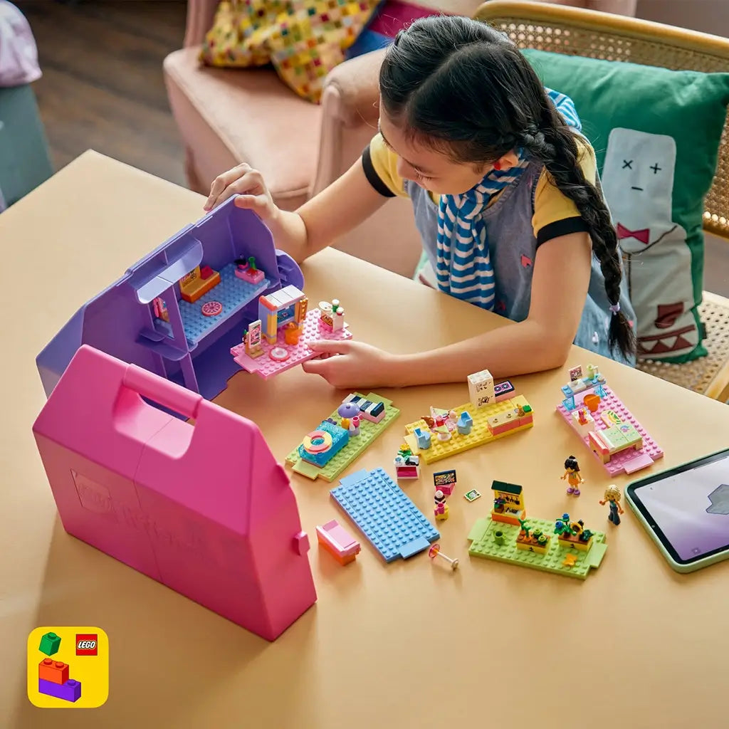LEGO Friends Creative Dollhouse Suitcase in pink and purple plastic sits open as a child builds colorful rooms and accessories at a table in a cozy home.