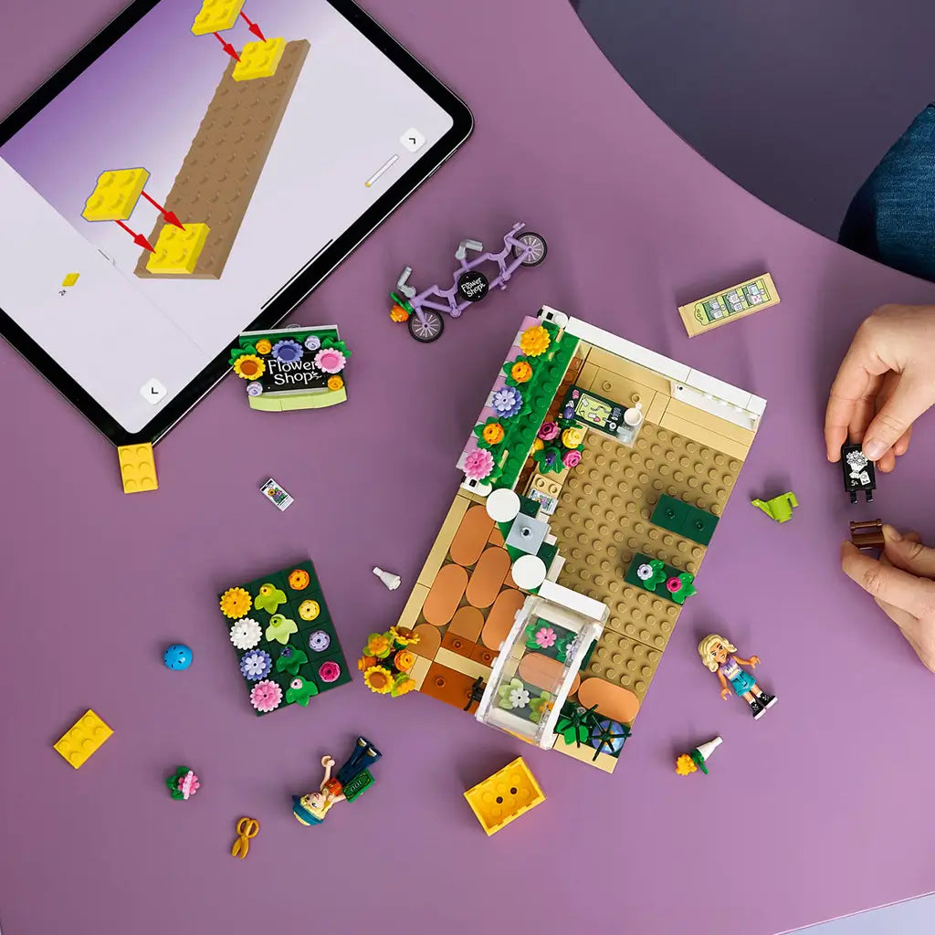 LEGO Friends Creative Flower Shop set in green, pink and white plastic, partly assembled with loose floral pieces beside digital building instructions on a tablet.