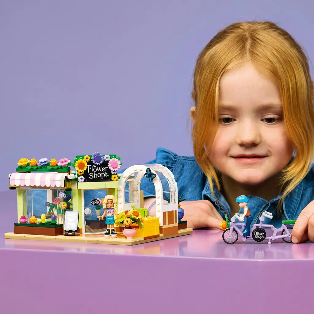 LEGO Friends Creative Flower Shop in green, pink and white plastic, with flower displays and tandem bicycle as a child plays with the set at a purple table.