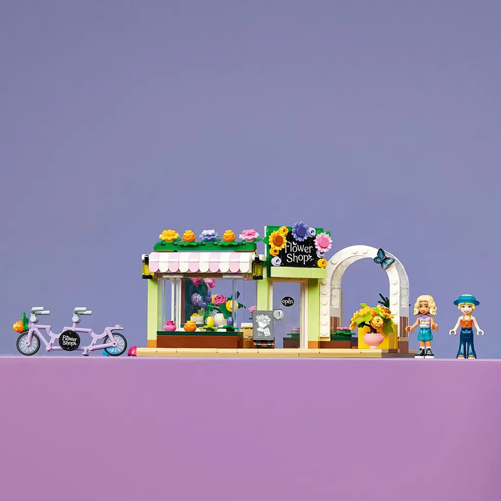 LEGO Friends Creative Flower Shop in green, pink and white plastic, with colorful blooms, tandem bicycle and two mini-dolls displayed on a purple tabletop.