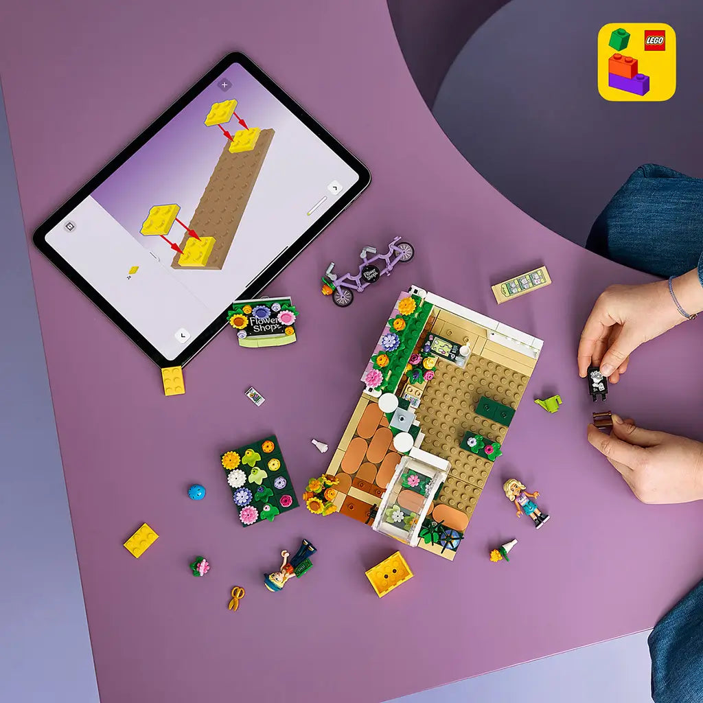 LEGO Friends Creative Flower Shop set in green, pink and white plastic, under construction with mini-dolls, bicycle and flower pieces beside digital instructions.
