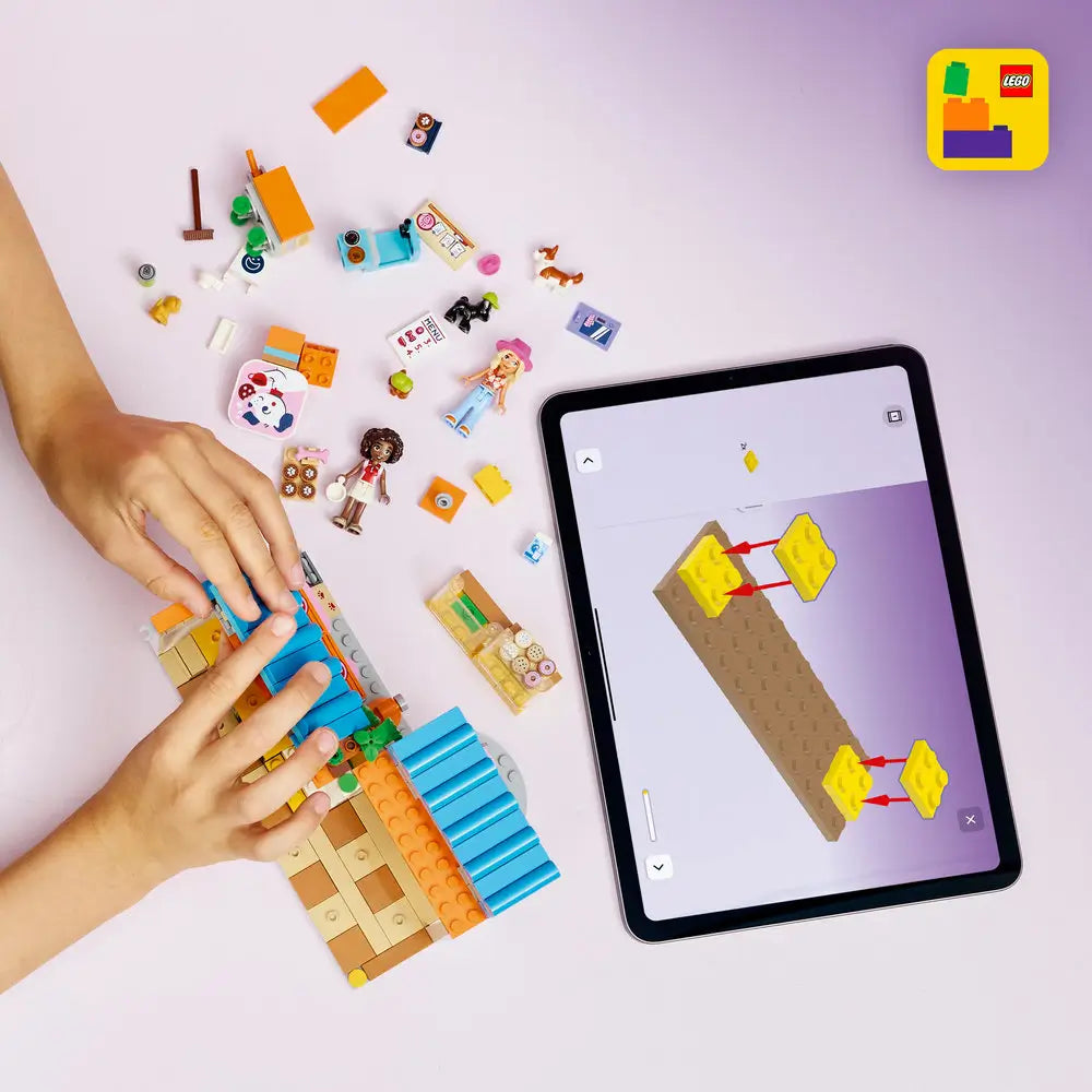 LEGO Friends building toy scene with dog bakery pieces spread on a light surface, showing a child assembling walls, blue roof panels, counters, treat accessories, pet figures, and minidolls using interactive digital building instructions.