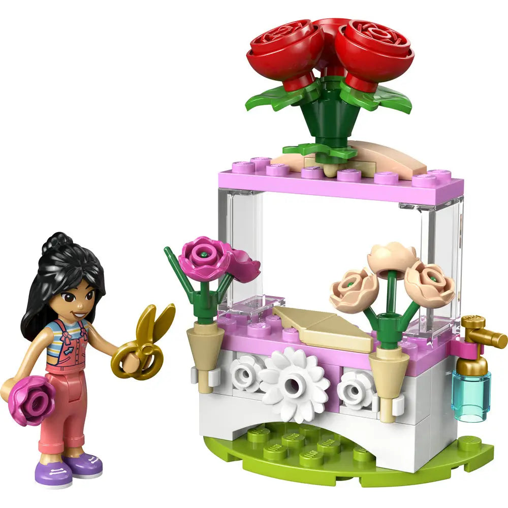 LEGO Friends flower stand with roses built from pink, white, green, and red plastic bricks, featuring a compact kiosk shape with clear panels, decorative flowers, watering tap, mini doll florist, and small garden accessories on a white studio background.