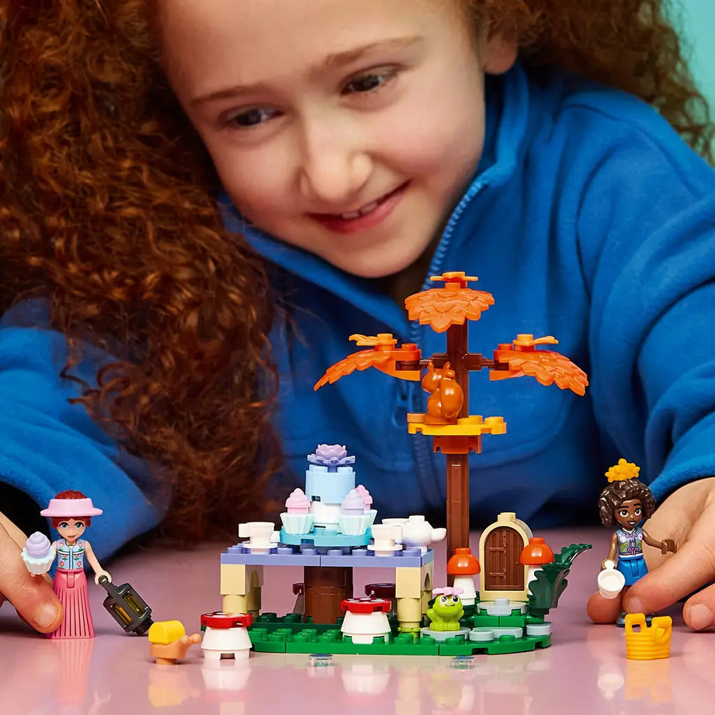 LEGO Friends Forest Tea Party set in green, brown and pastel plastic, with tea table, autumn tree and animal figures as a child plays with the scene on a pink table.