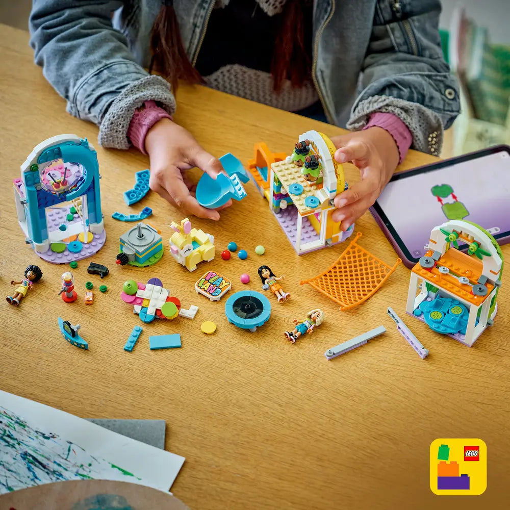 LEGO Friends building toy scene with colorful plastic playground pieces spread on a wooden table, showing a child assembling arches, slide elements, figures, balls, and accessories using digital guided instructions.