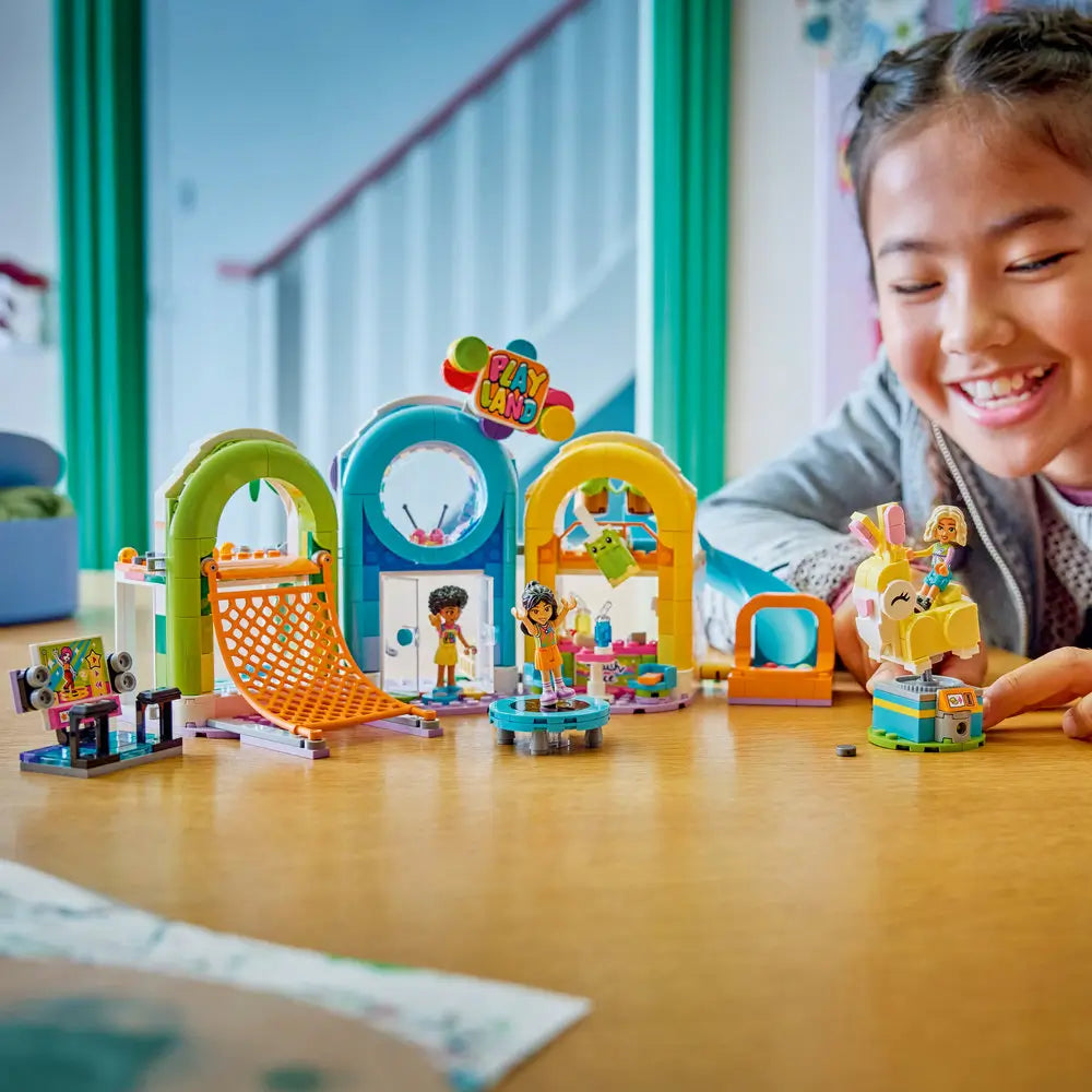 LEGO Friends playset lifestyle scene with a smiling child interacting with a completed indoor playground, moving minifigures between the slide, trampoline, snack counter, and ride-on toy during creative play.