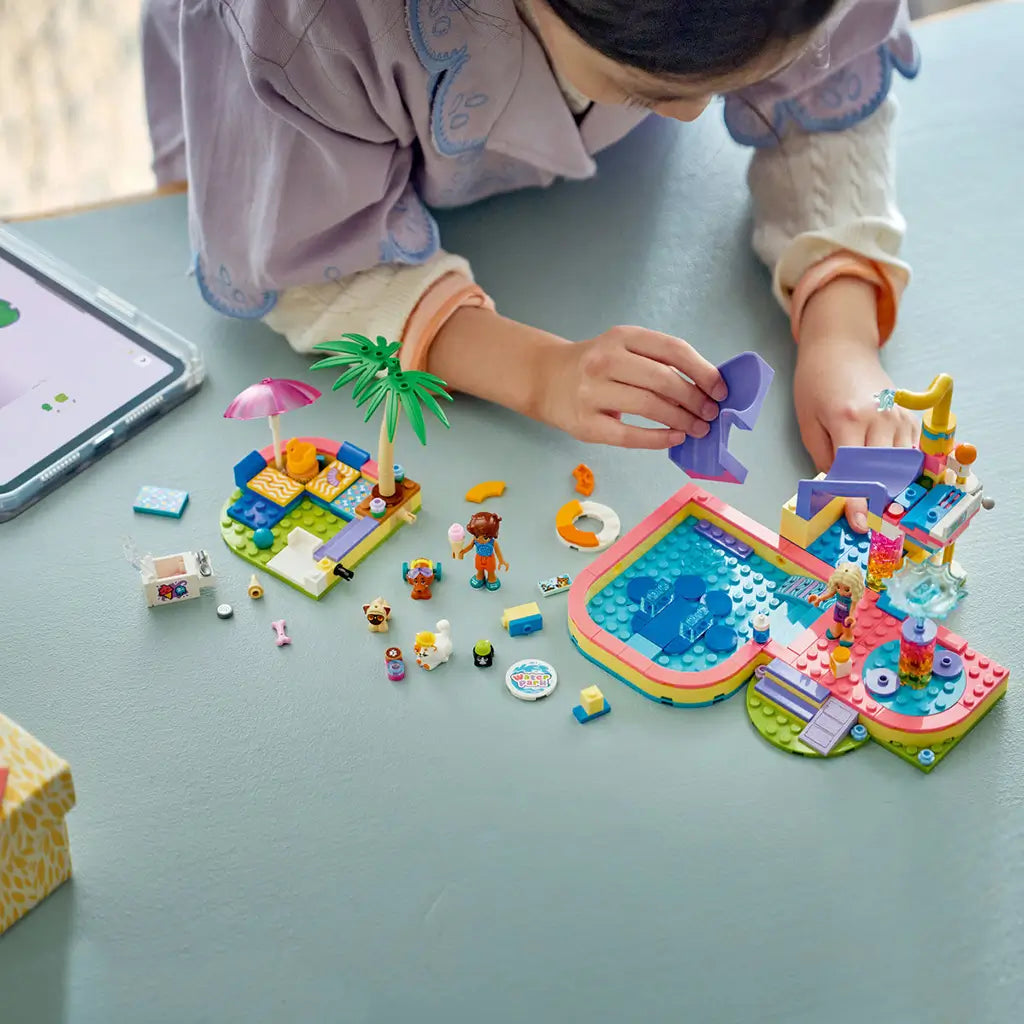 LEGO Friends Fun Water Park Day build in pink, aqua and purple plastic, with a child attaching the slide beside the pool, mini-dolls, pets and loose toy pieces.