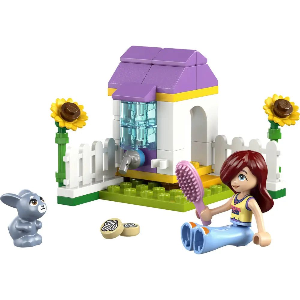 LEGO Friends garden bunny house playset built from pastel purple, white, yellow, and green plastic bricks, featuring a small arched shelter with water dispenser, fenced base, sunflower details, a bunny figure, and a seated mini doll on a white studio background.