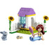 LEGO Friends garden bunny house playset built from pastel purple, white, yellow, and green plastic bricks, featuring a small arched shelter with water dispenser, fenced base, sunflower details, a bunny figure, and a seated mini doll on a white studio background.