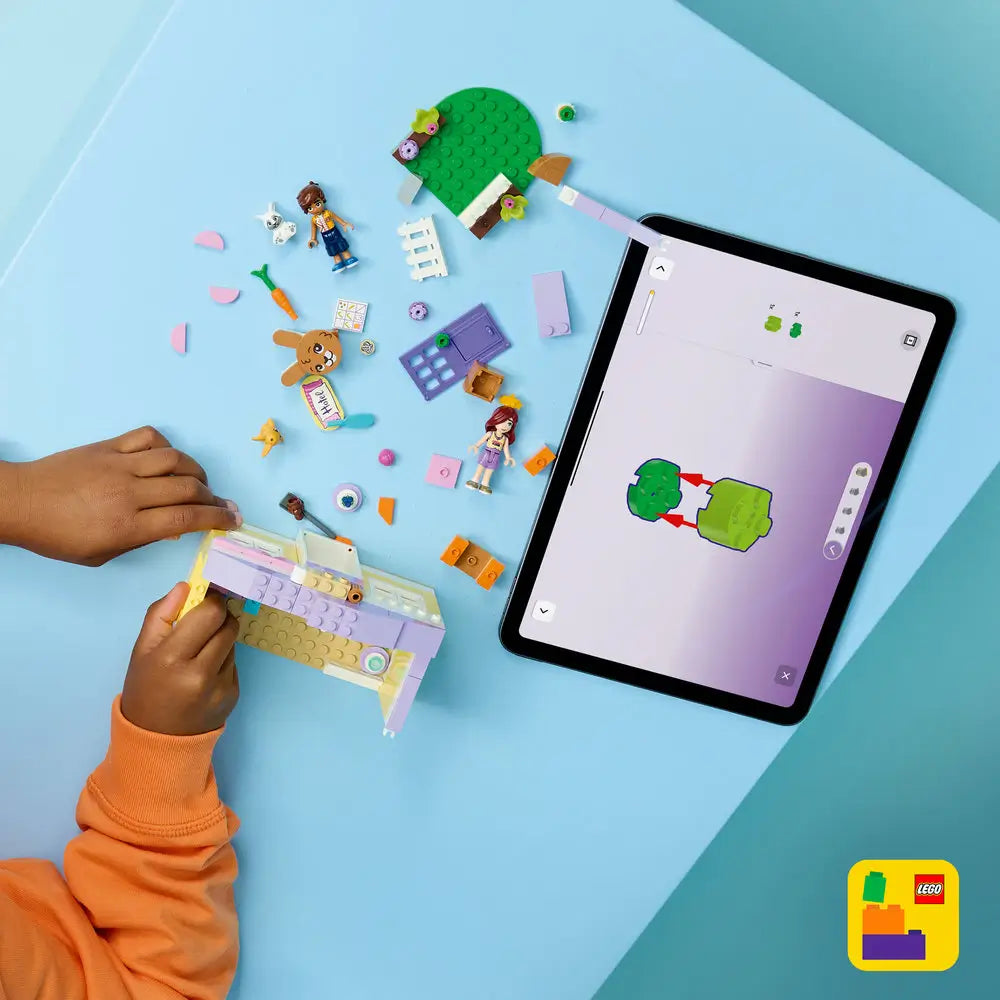 LEGO Friends building toy scene with pastel hotel pieces spread on a blue surface, showing a child assembling the bunny hotel walls, roof, garden base, animals, and minifigures using digital building instructions.