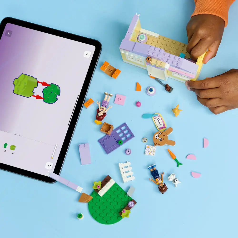 LEGO Friends lifestyle image showing a child building a bunny hotel from pastel plastic bricks, connecting walls and roof sections while animals, plants, and accessories are arranged for imaginative play.