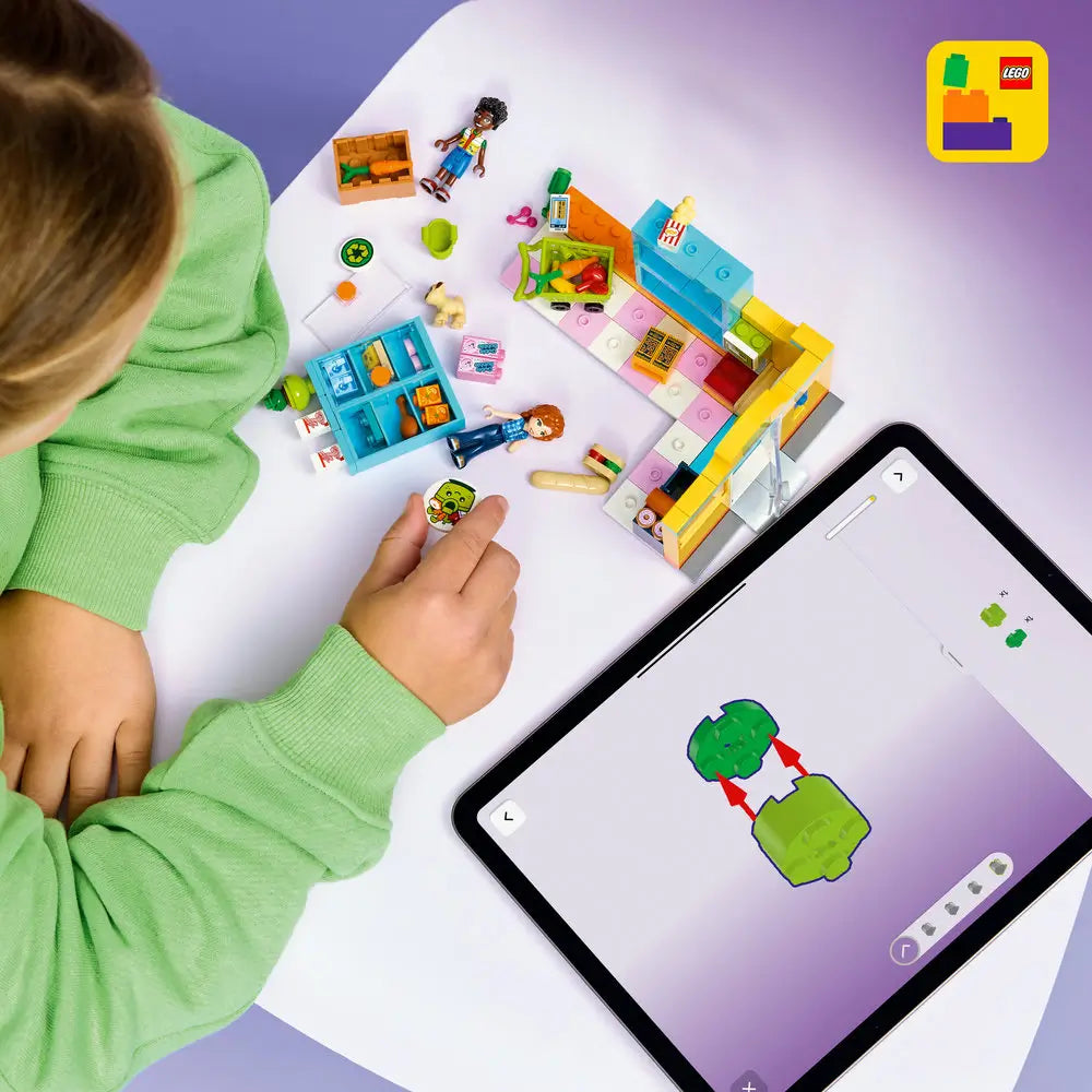 LEGO Friends building toy scene with colorful store pieces spread on a white table, showing a child assembling shelves, checkout counter, food accessories, minifigures, and a shopping cart using digital guided instructions.