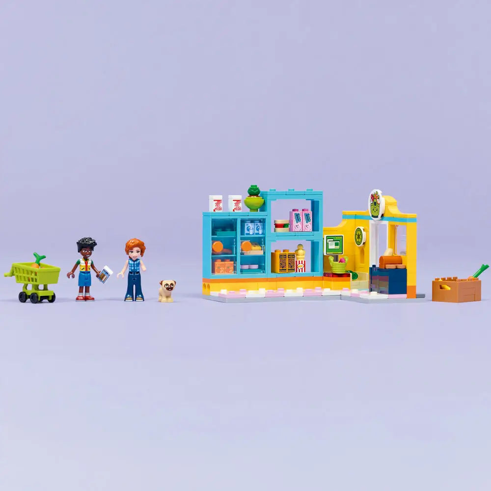 LEGO Friends convenience store display showing teal and yellow plastic bricks forming a finished grocery shop with stocked shelves, checkout area, recycling sign, shopping cart, dog figure, and minifigures posed.