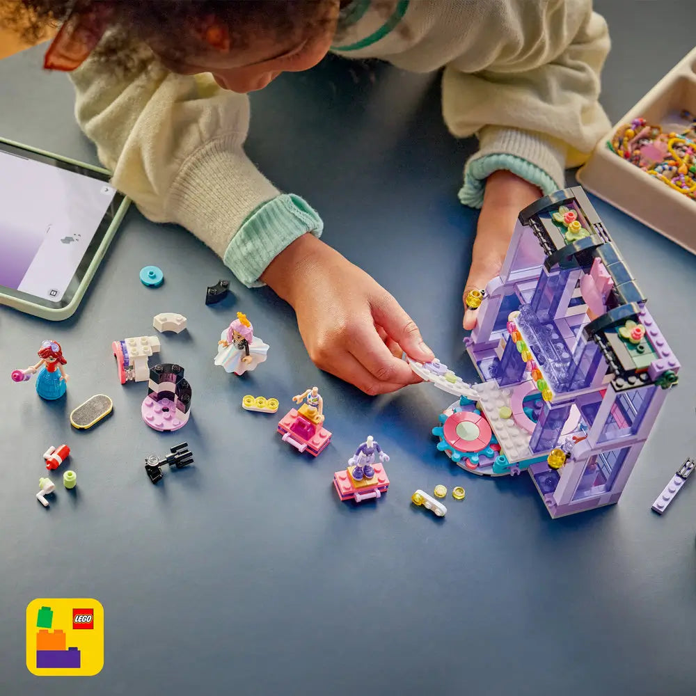 LEGO Friends building toy scene showing pastel purple plastic bricks spread on a tabletop, with a child assembling a fashion show runway, stage platform, figures, outfits, and accessories using digital guided instructions.