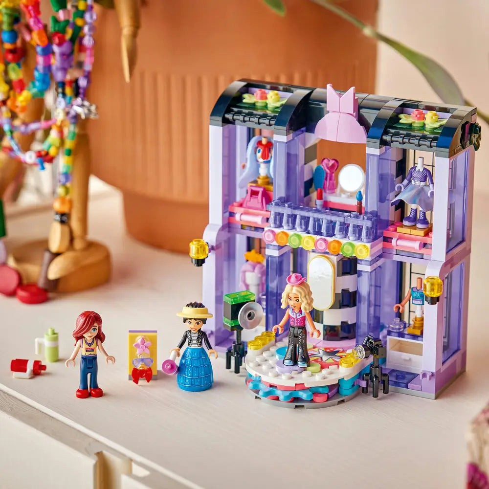 LEGO Friends fashion show display showing lavender and purple plastic bricks forming a finished runway building with rotating stage, balcony, lights, outfit displays, and multiple minifigures posed on a tabletop.