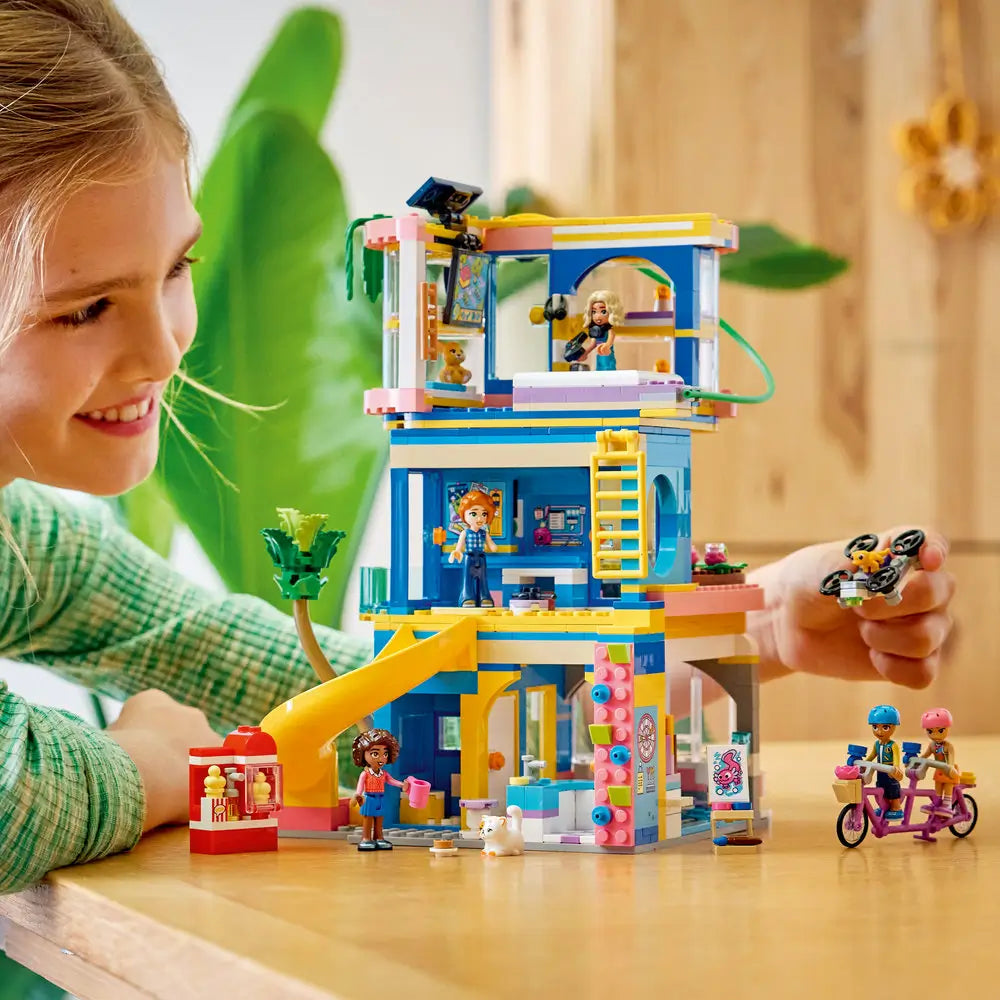 LEGO Friends Heartlake City Friends Club House lifestyle image shows a child building and playing with the blue and pink plastic club house, arranging mini dolls, slide, café items, and tech accessories on a tabletop.